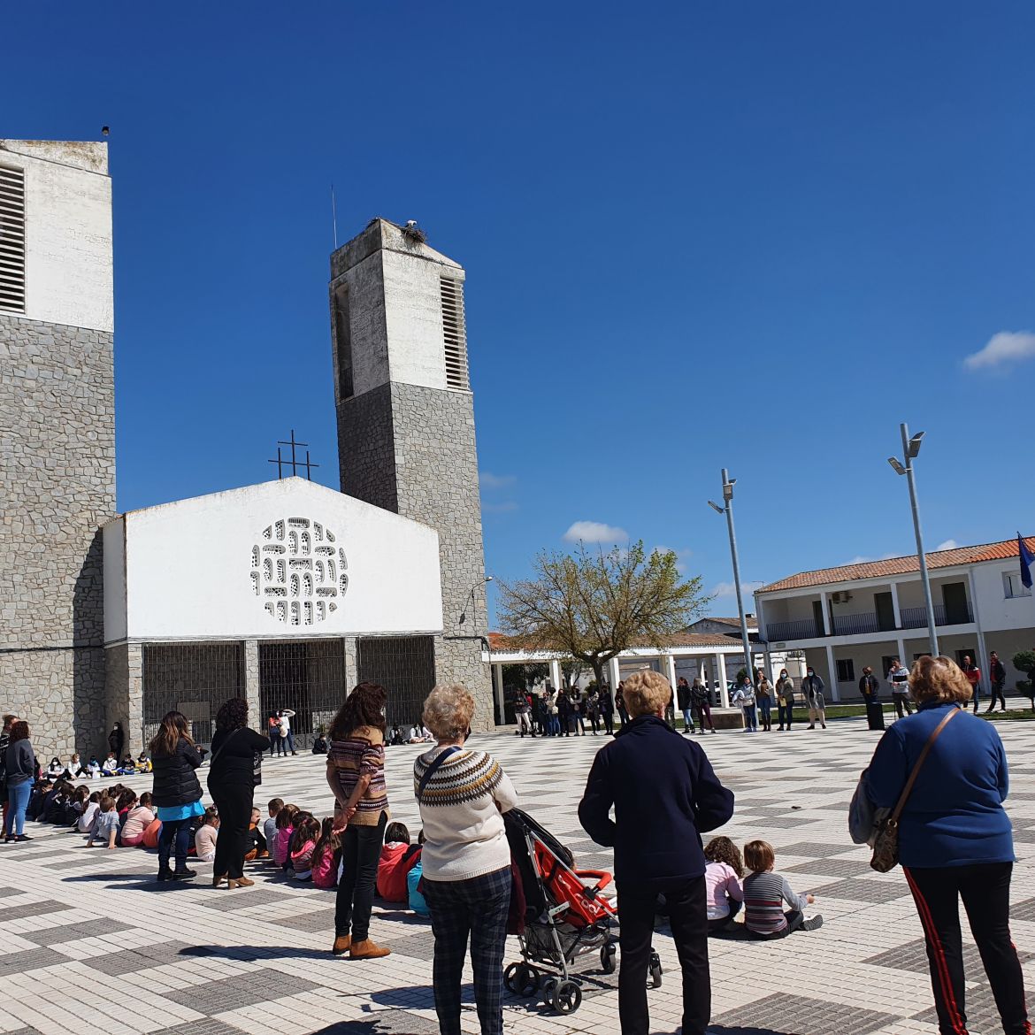 Plaza Principal del municipio Hernán Cortés delimitada por la Iglesia y el Ayuntamiento. Niños sentados en línea acompañados por adultos.