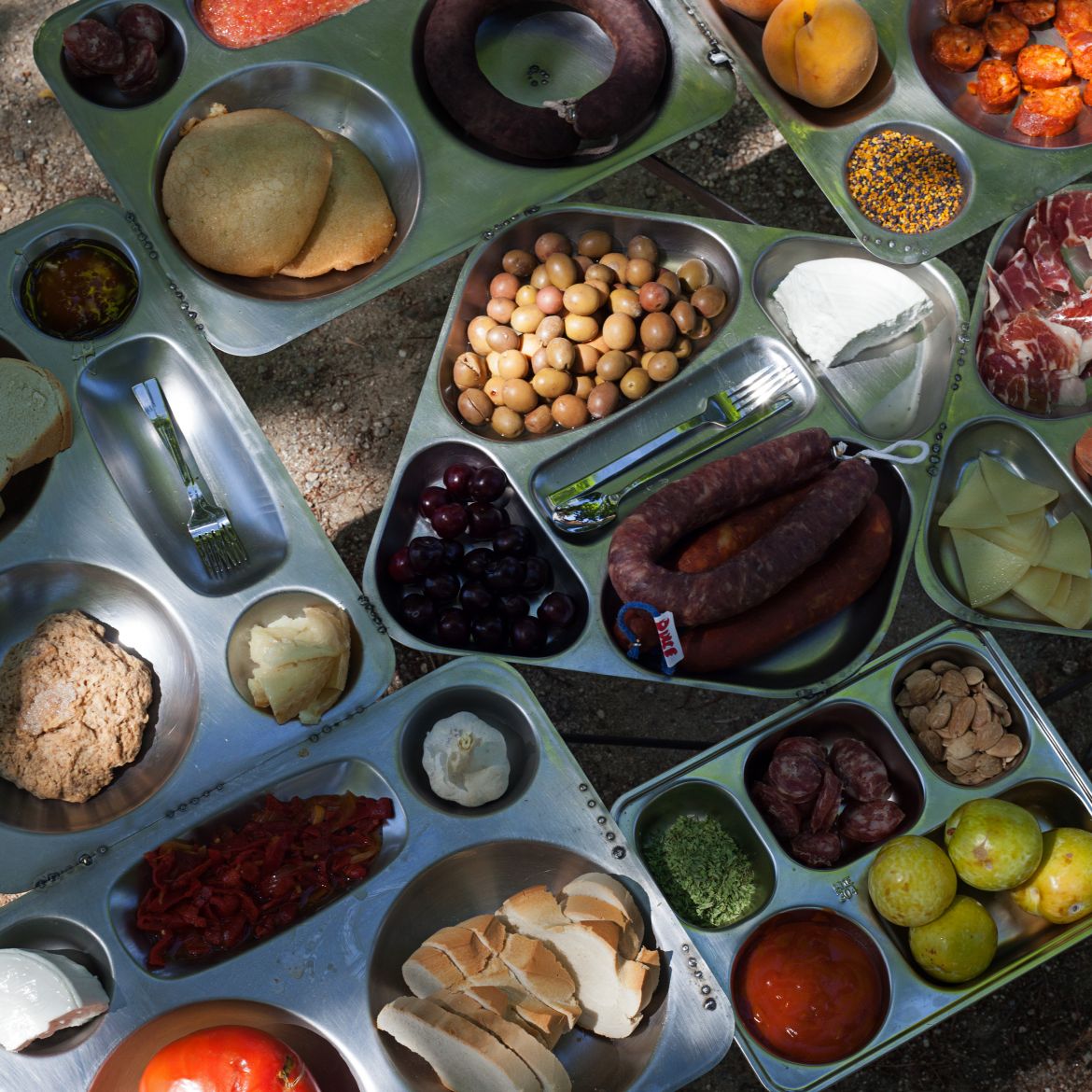 Bandejas metálicas llenas de comida: Pan, queso, cabeza de ajo, cubiertos, aceitunas, tomate, melocotón, embutido