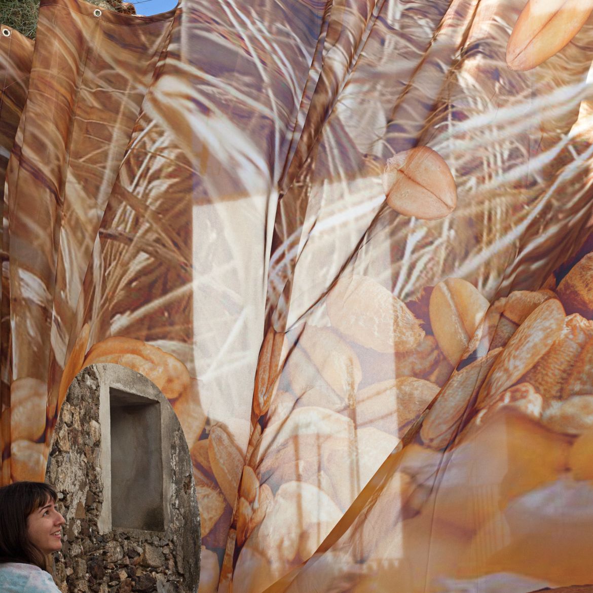 esquina inferior izquierda, mujer mirando hay un lado de la calle. Segundo plano: Textil translucido con motivos vegetales. deja entrever fachada de piedra.